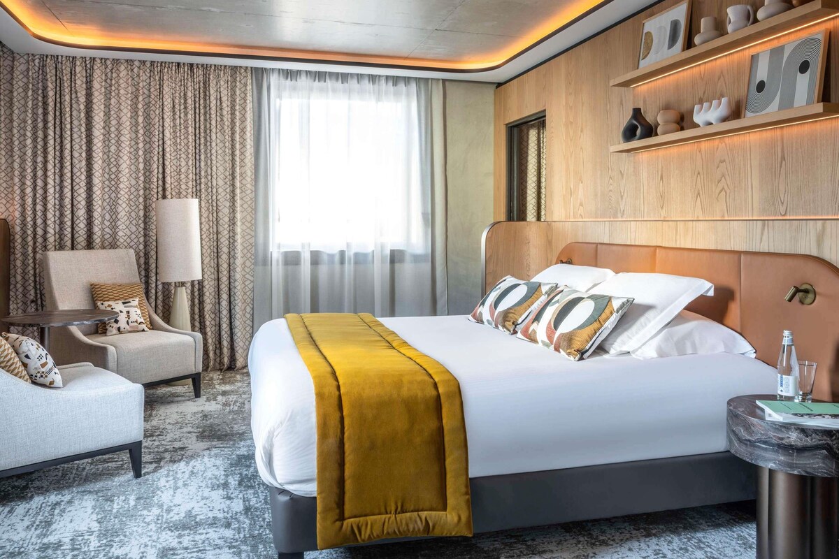 A modern hotel room with a large bed, a seating area with a chair and a table, and a shelf with decorative items.
