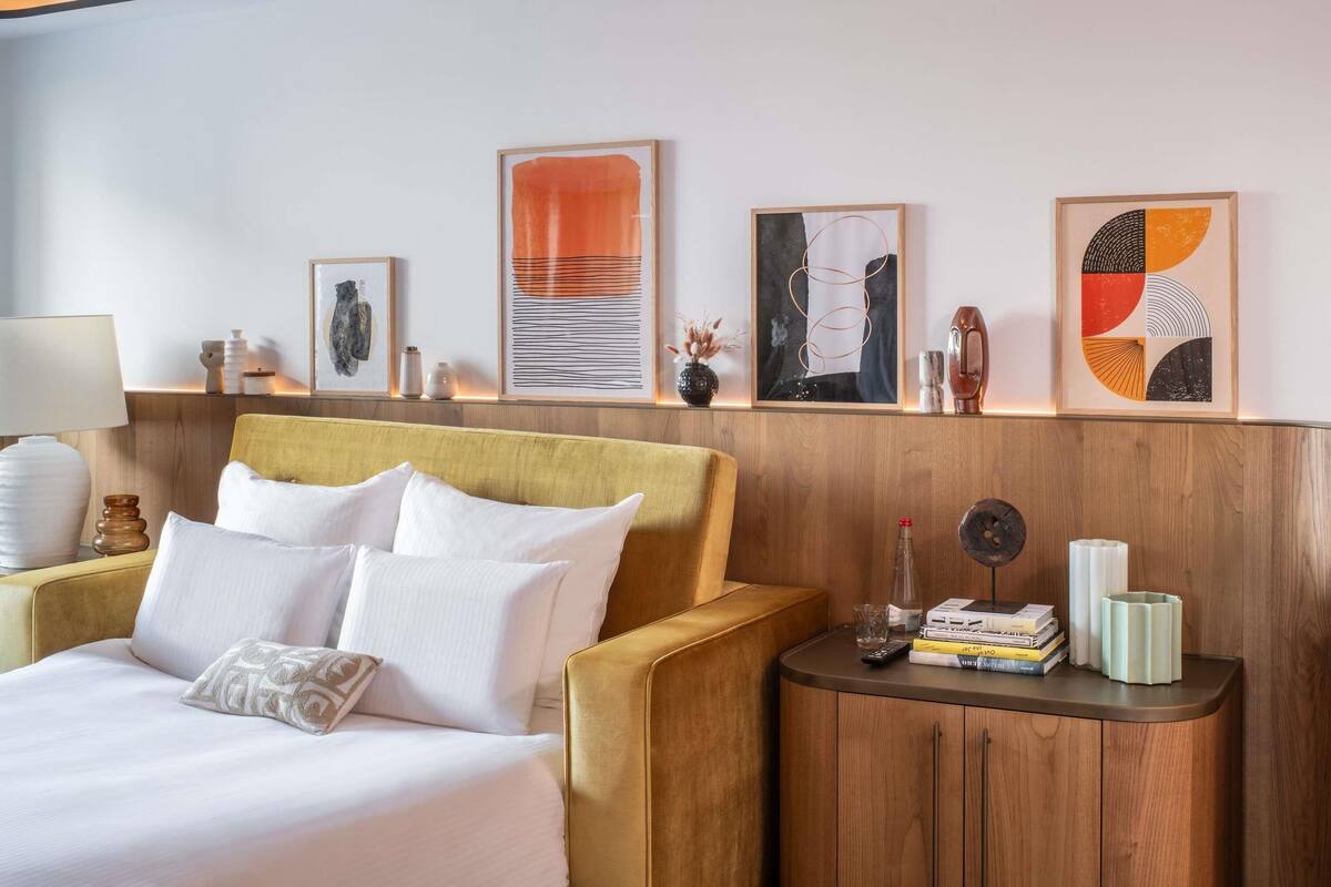 A hotel room with a bed, a sofa, a nightstand with books and a remote control, and artwork on the wall.