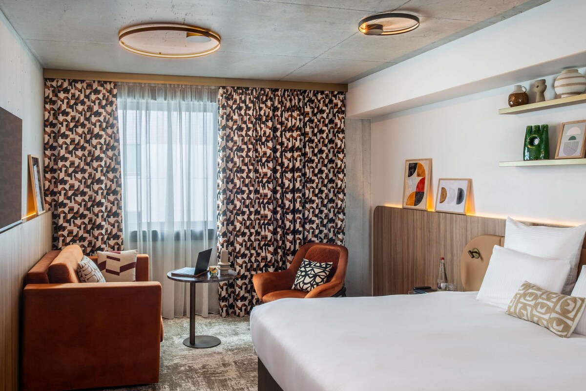 A hotel room with a bed, a sofa, a chair, a small table, and a window with curtains.