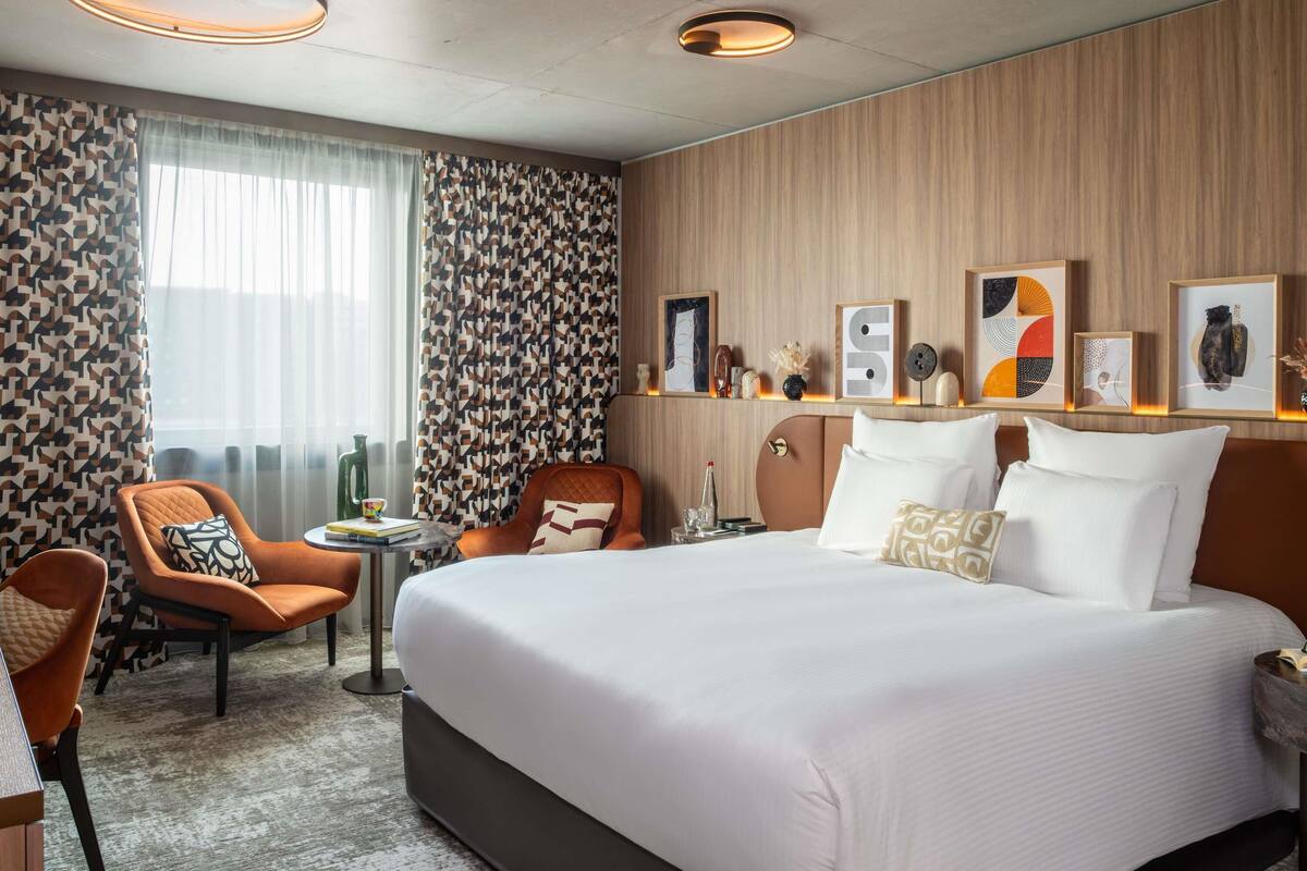 Een hotelkamer met een groot bed, twee fauteuils, een tafeltje en een raam met gordijnen.