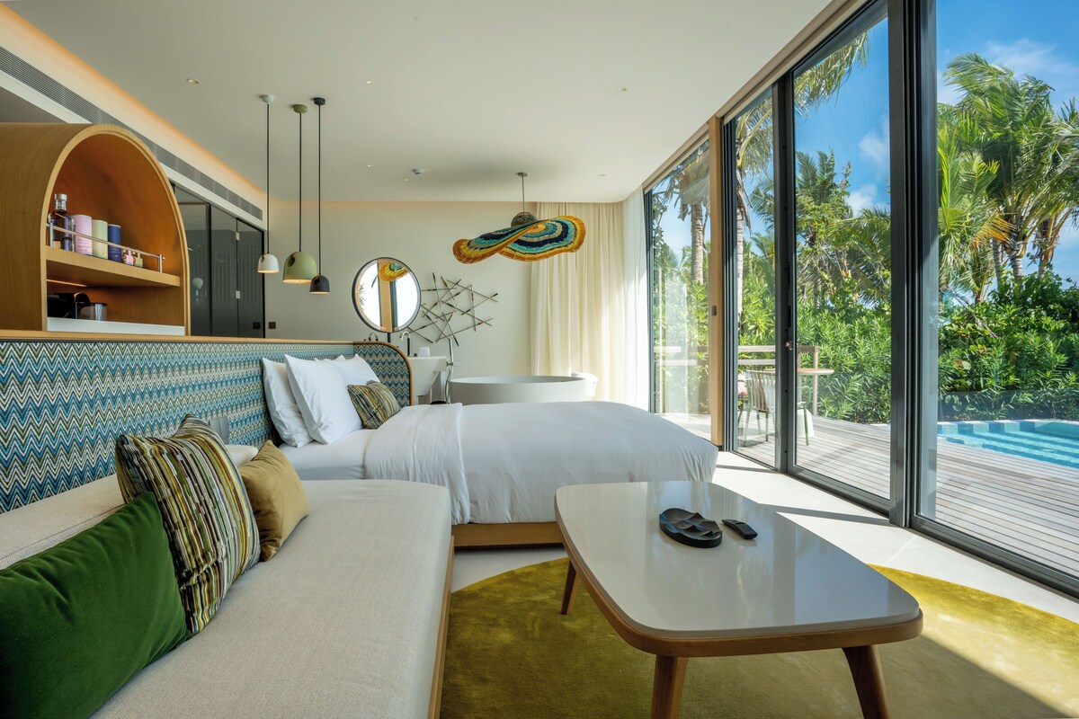 A modern hotel room with a large bed, a sofa, a dining table, and a view of a pool and tropical landscape.