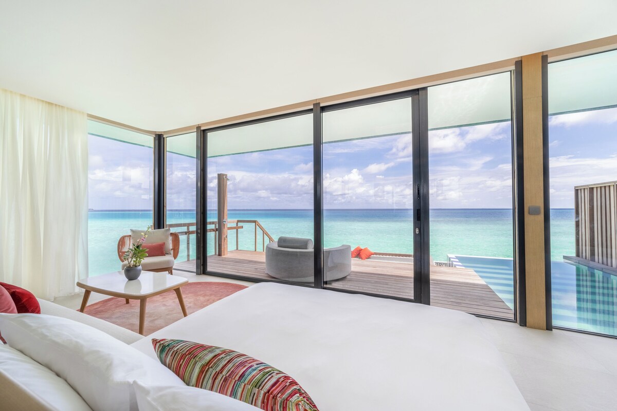 A modern hotel room with a large bed, a seating area with a view of the ocean, and a balcony with a pool.