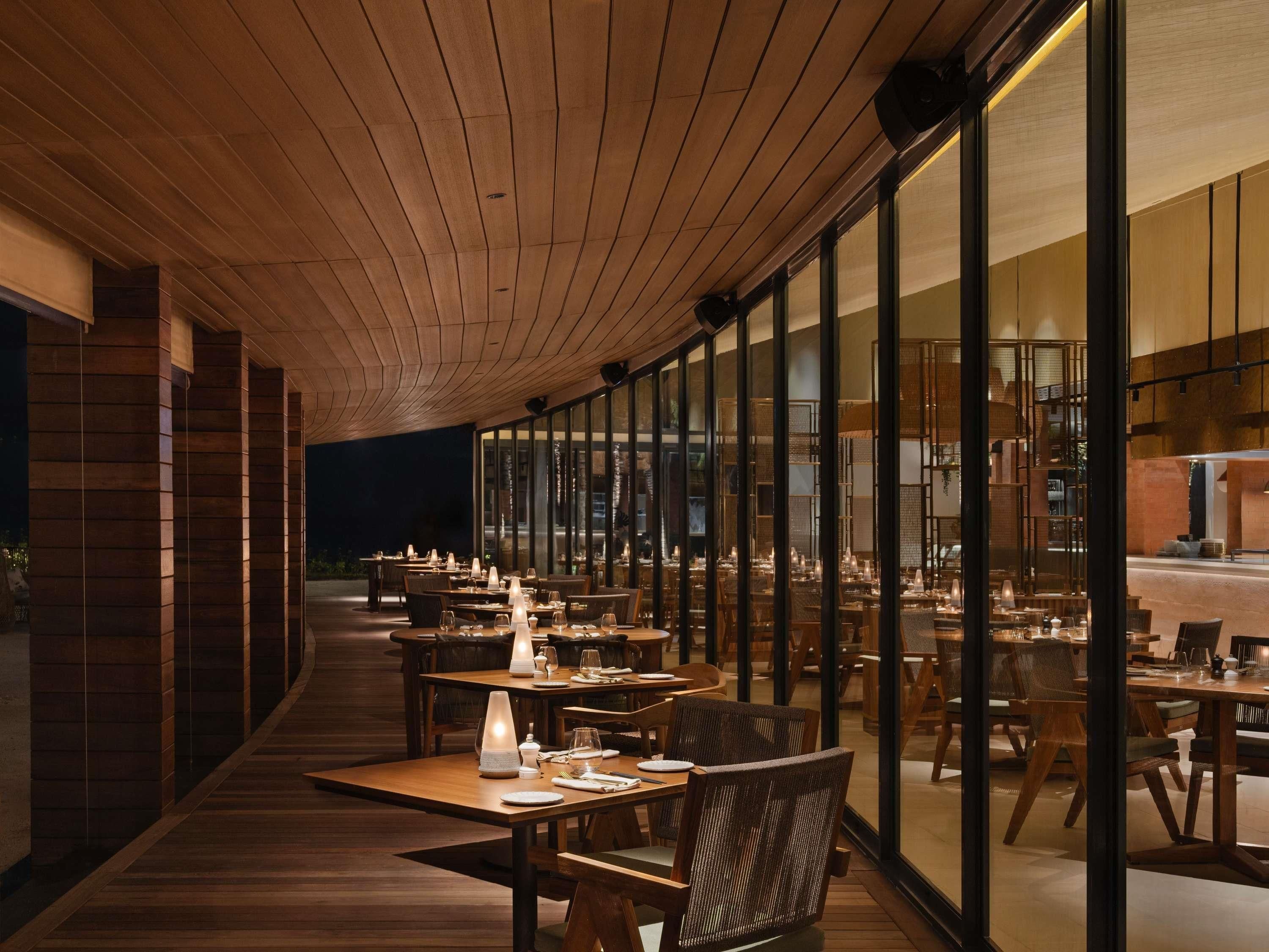 Un'area pranzo interna con tavoli e sedie in legno, ampie finestre e soffitto in legno.