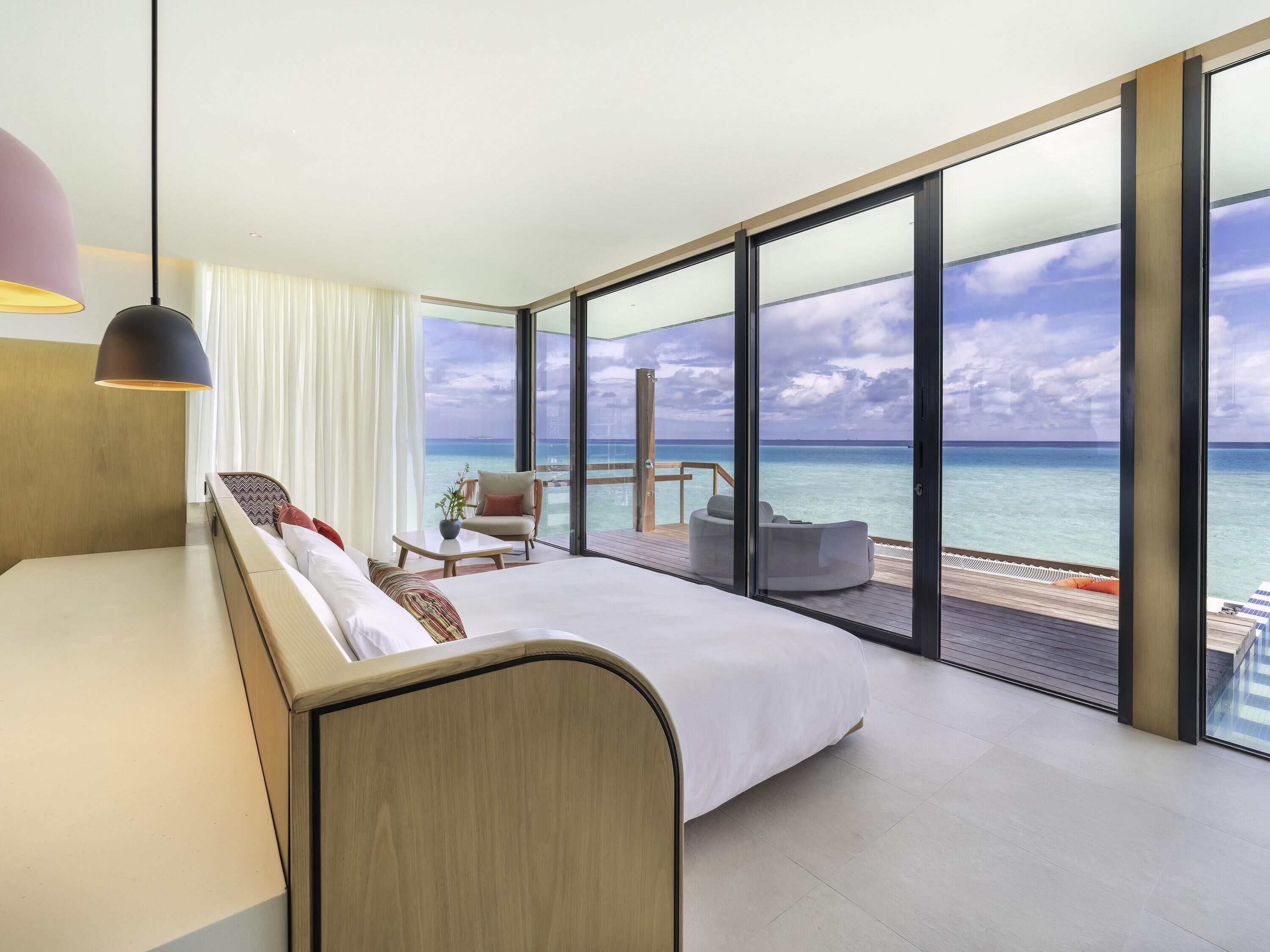 A modern hotel room with a large bed, a seating area with a view of the ocean, and a balcony with a wooden deck.