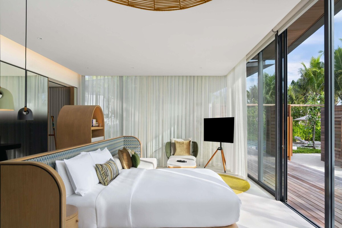 A modern bedroom with a large bed, bedside tables, a TV on a tripod, and a view of the outdoors.