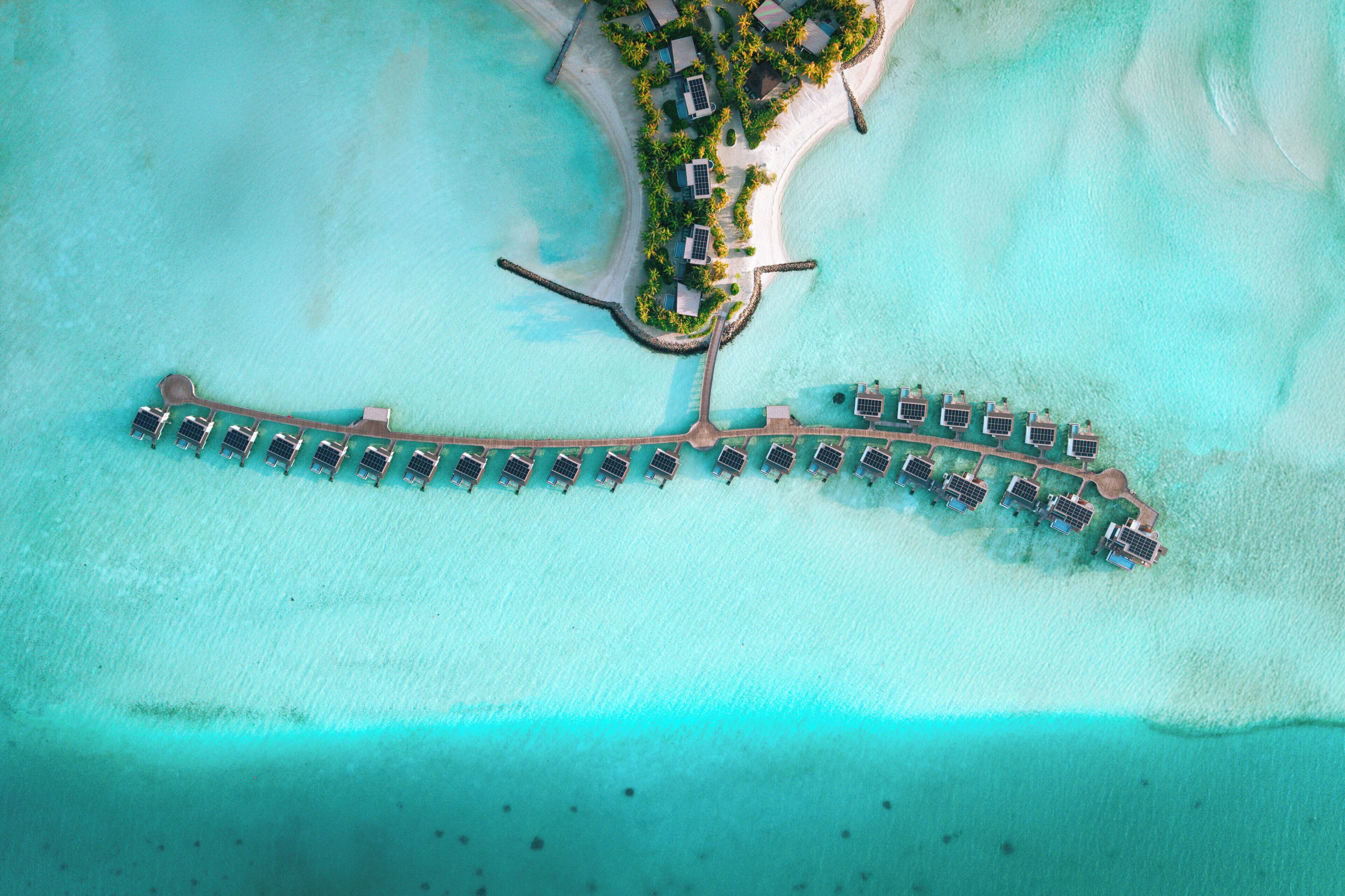 Vista aérea de un complejo turístico con un muelle largo que conduce a una fila de bungalows sobre el agua, unidos por un puente.
