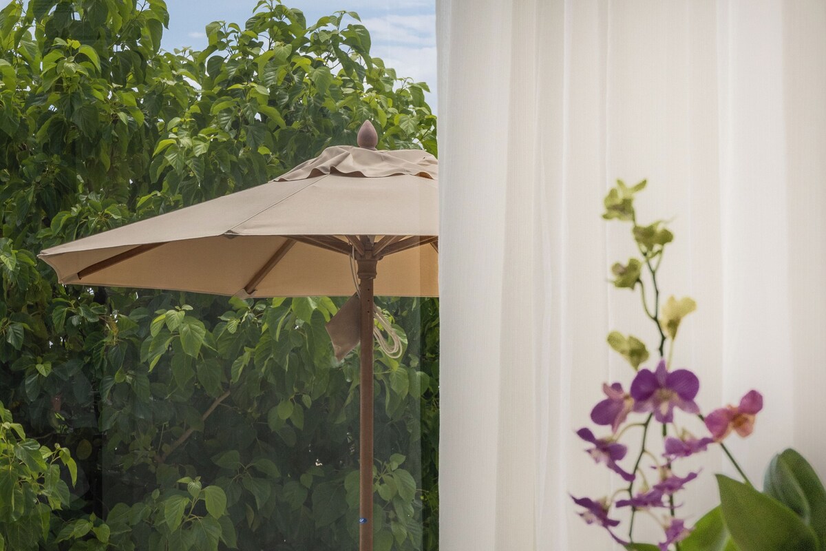 A sunlit room with a beige sofa, a striped pillow, and a green pillow. An umbrella is partially visible, and there is a potted plant with purple flowers.