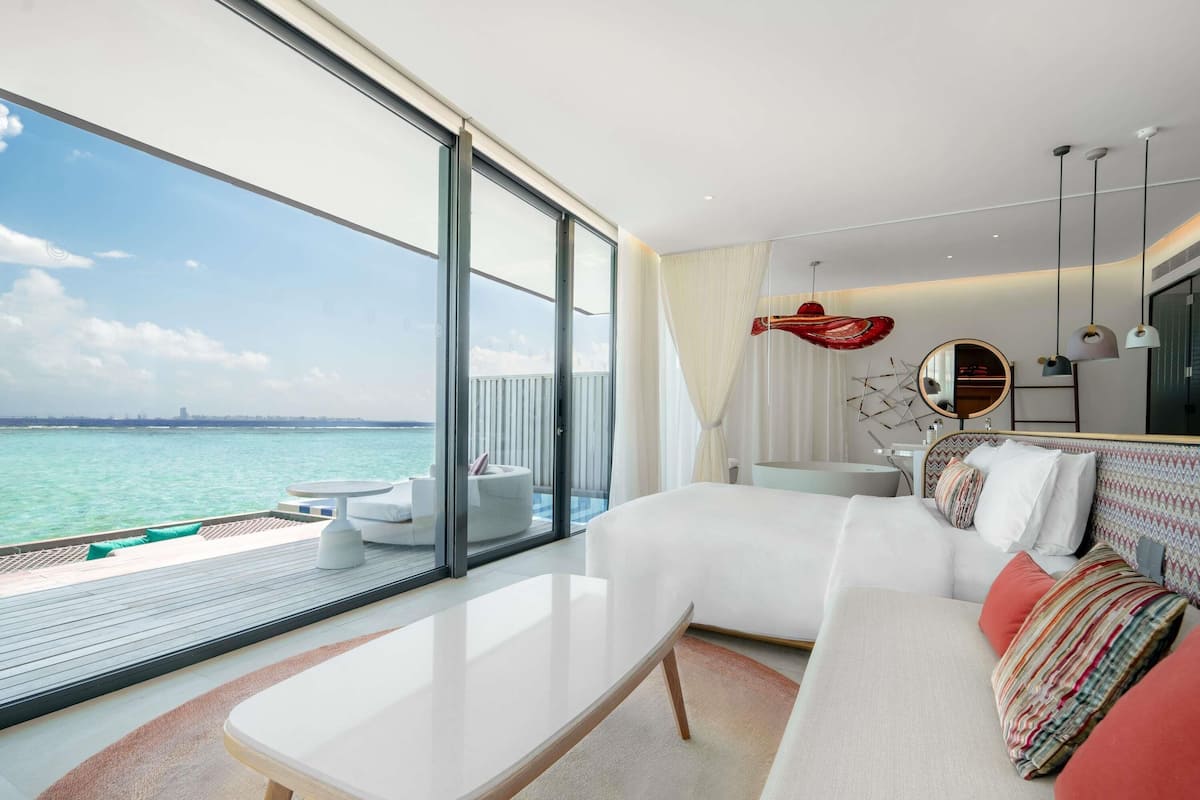 A modern hotel room with a large balcony, a bed, a small table, and a view of the sea.