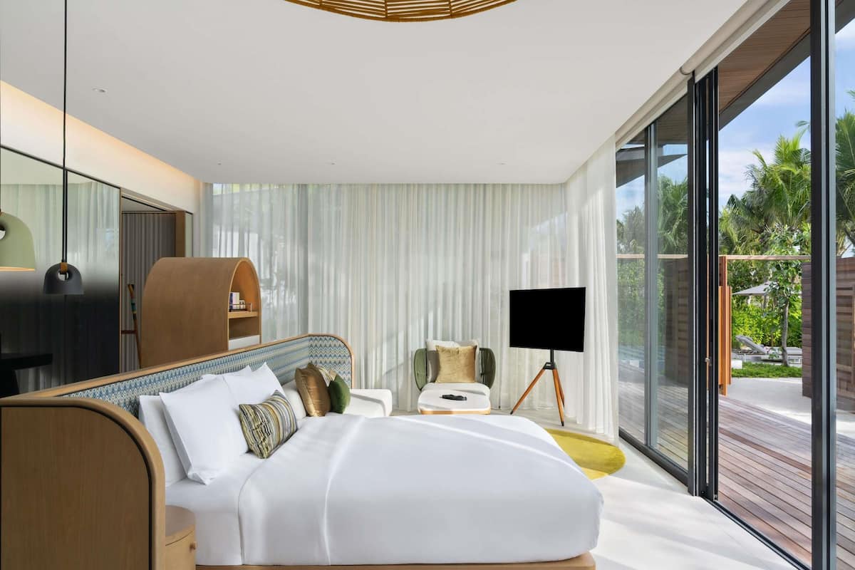 A modern bedroom with a large bed, bedside tables, a TV on a tripod, and a view of the outdoors.