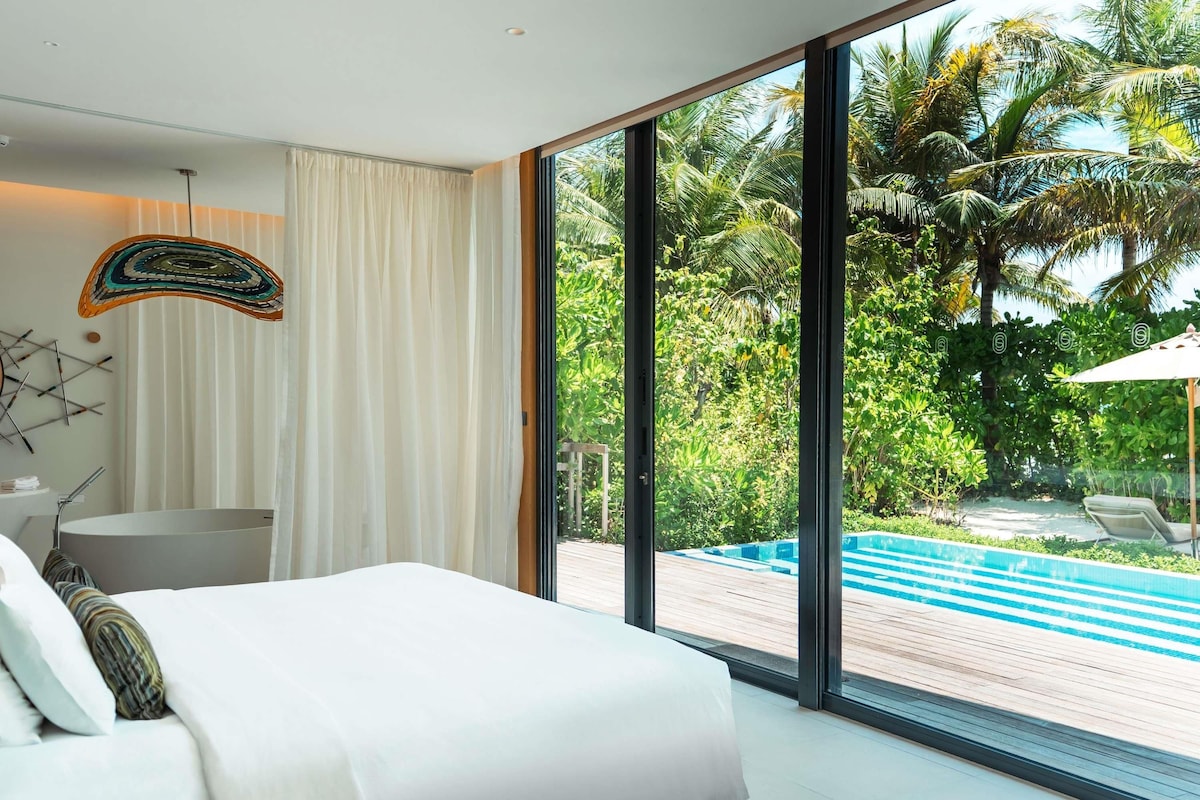 A modern hotel room with a large bed, a view of a pool, and a tropical garden.
