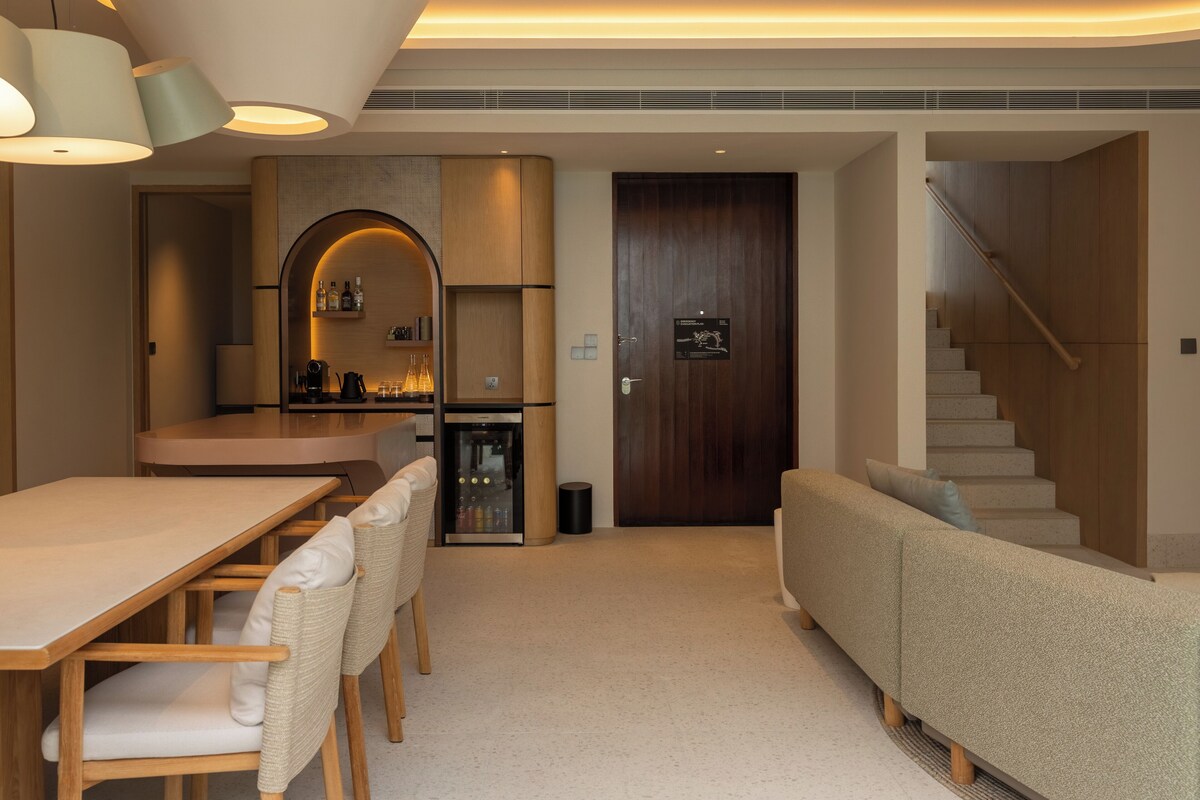 A modern living room with a wooden dining table, cushioned chairs, a sofa, and a built-in kitchen area with a refrigerator and wine cooler.