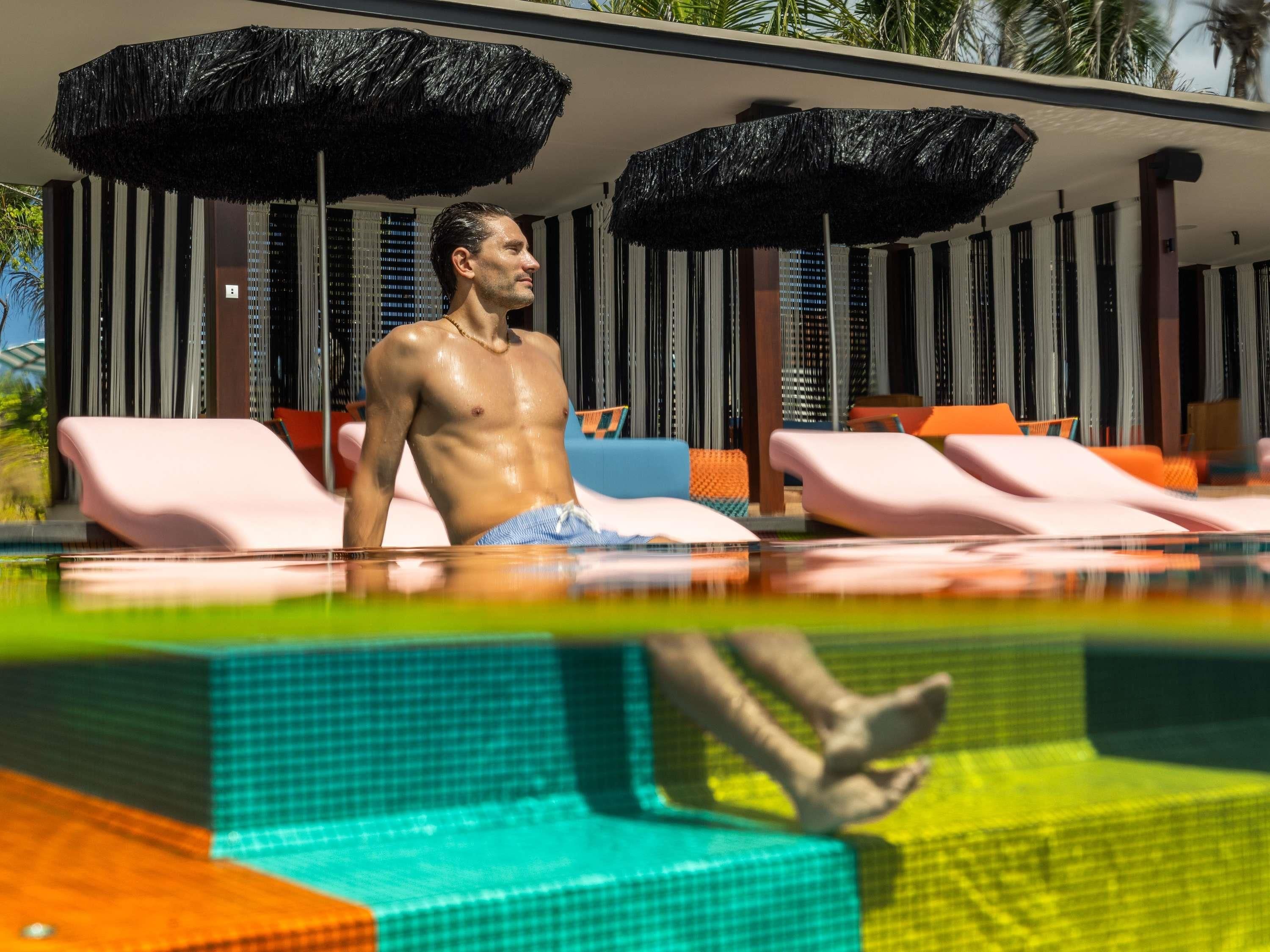 A man in a swimming pool with lounge chairs and black umbrellas.