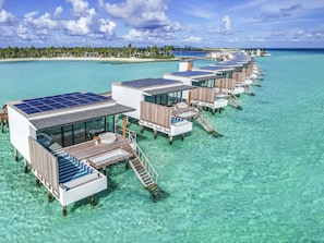 Cabañas sobre el agua con paneles solares, agua turquesa cristalina y una playa al fondo.