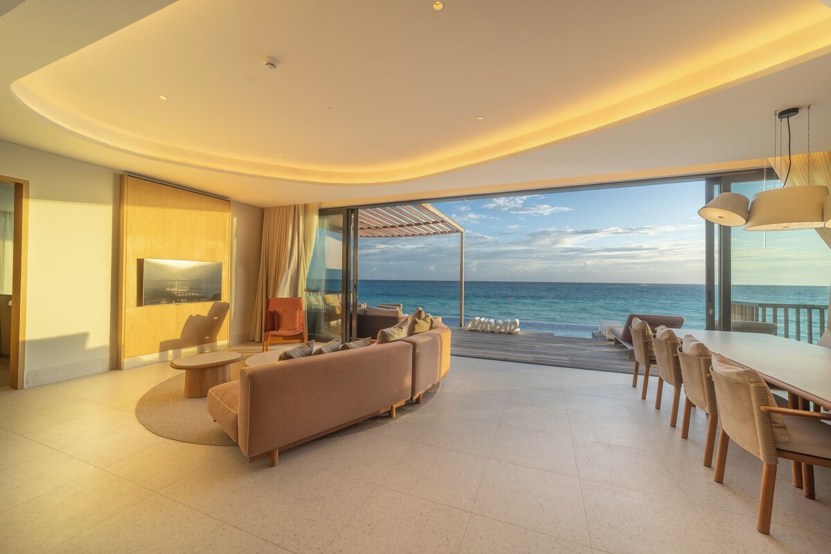 A modern living room with a large window overlooking the ocean, a sofa, a dining table, and a TV.