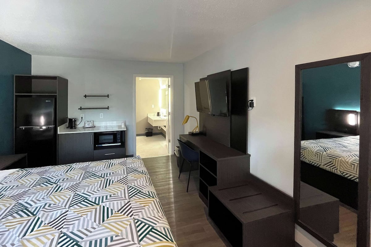 A hotel room with a bed, a desk, a TV, a mirror, and a kitchenette.