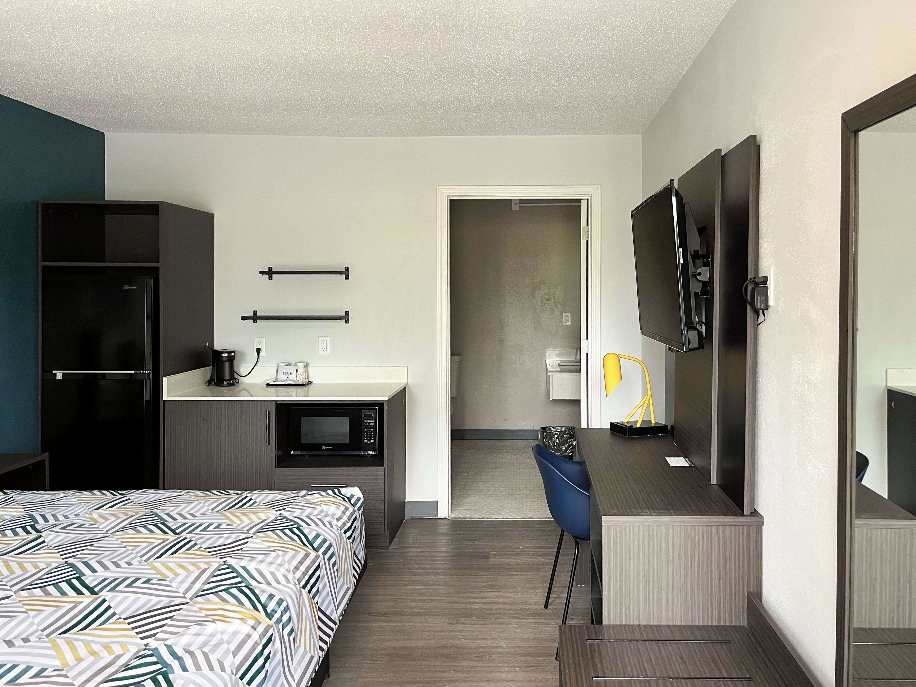 A hotel room with a bed, a desk with a microwave, a refrigerator, a TV, and a bathroom visible through an open door.