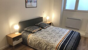 Ein ordentlich bezogenes Bett mit gemusterten Betttextilien, zwei Nachttische mit Lampen, ein gerahmtes Bild an der Wand und ein Fenster mit Jalousien.