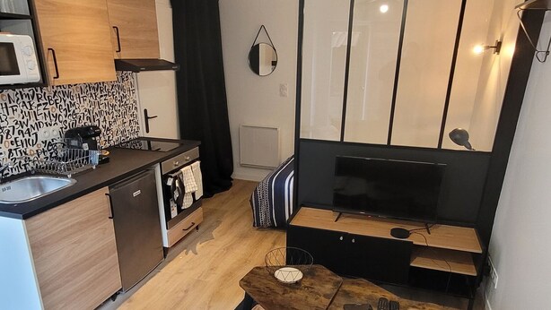 A modern living room with a grey sofa, a coffee table, and a TV. There's a kitchen area with wooden cabinets and a microwave. - Studio cosy Saint-Étienne centre (Saint-Ã‰tienne)