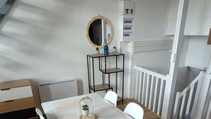 Un área de comedor moderna con una mesa y sillas blancas, una mesita auxiliar con objetos decorativos y una escalera al fondo.