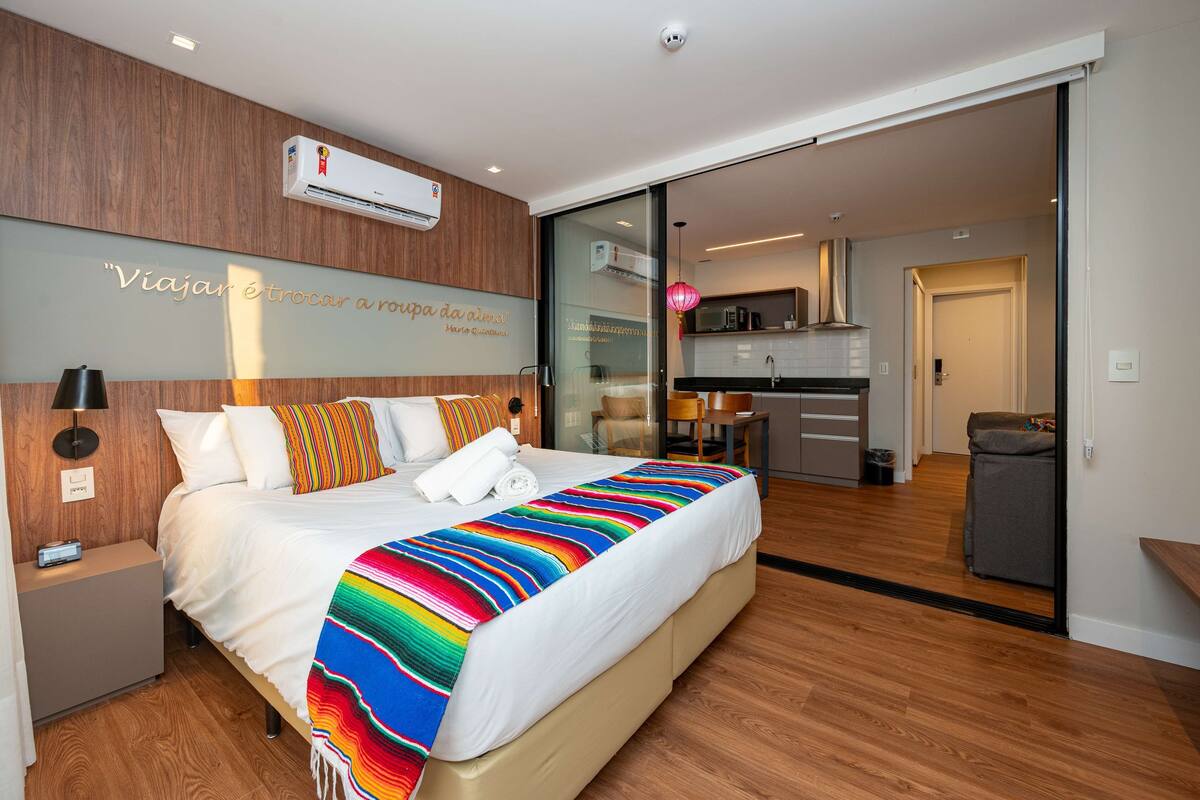 Una habitación de hotel moderna con una cama grande, una mesita de noche, aire acondicionado montado en la pared y una cocina visible a través de una puerta de vidrio.