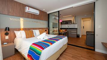 Una habitación de hotel moderna con una cama grande, una mesita de noche, aire acondicionado montado en la pared y una cocina visible a través de una puerta de vidrio.