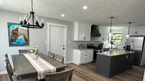 A modern kitchen with white cabinetry, a central island, and a dining area with a wooden table and chairs. - 🐎 2 Miles to Flintfields, Fenced Yard pet friendly, FREE RENTAL Golf Cart (Williamsburg)