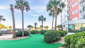 A landscaped area with palm trees and green turf, surrounded by a modern building with large windows. - NEW! Palace Resort TRUE Oceanfront STUDIO Condo (Myrtle Beach)