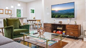 A modern living room with a green armchair, a glass coffee table, a wooden cabinet, and a flat-screen TV displaying a scenic landscape. - Fall Getaway in Asheville w/Hot Tub & Fire Pit! (Asheville)