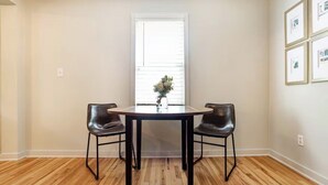 A dining area with a round table, two chairs, a vase with flowers, and a window with blinds. - Charming Park Circle Bungalow with Screened Porch (North Charleston)