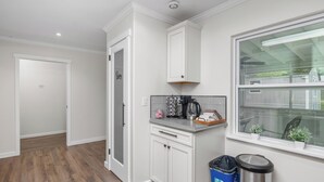Een keuken met witte kastjes, een aanrecht, een raam met planten en een prullenbak.