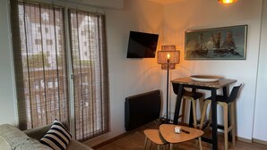 A living room with a sofa, a dining table with chairs, a TV mounted on the wall, and a painting above the table. - Joli Studio Avec Balcon en Bord de mer (Cabourg)
