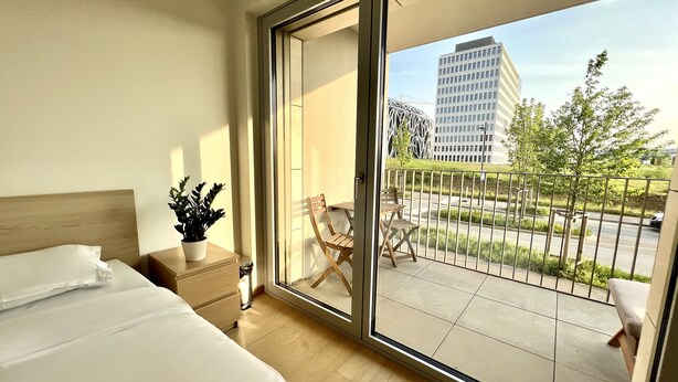Habitación de hotel con cama, mesita de noche con planta y puerta corredera de vidrio que da a un balcón exterior con vistas a un paisaje urbano y un edificio de fachada singular.
