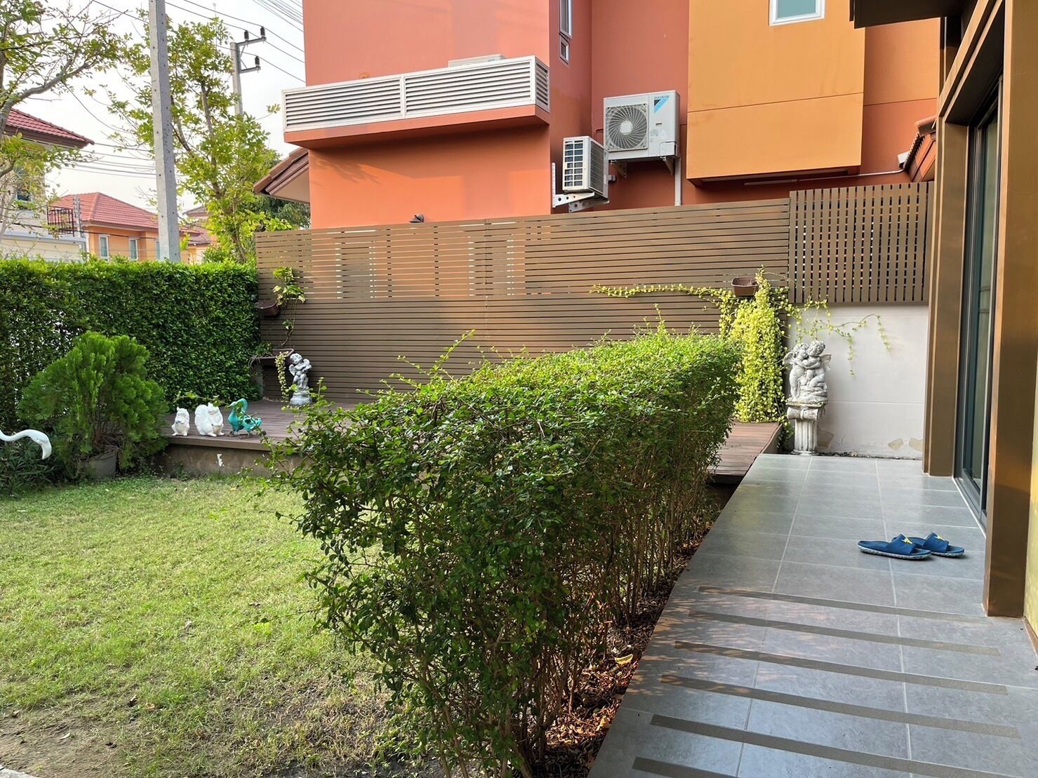 A residential area with a well-maintained garden, a wooden fence, and a pathway leading to a building with air conditioning units.