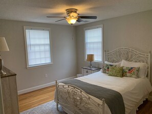 Un dormitorio con cama blanca, mesita de noche, lámpara y ventilador de techo.
