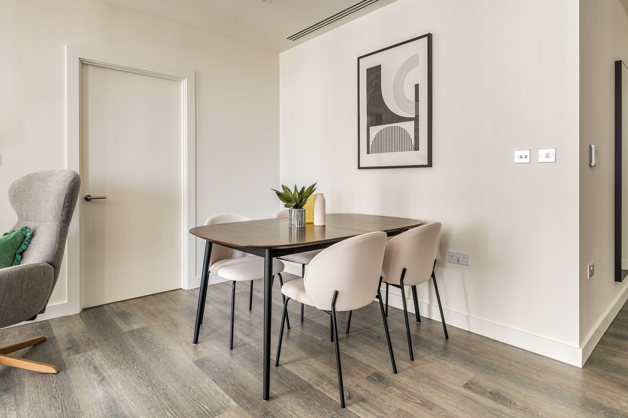 A dining area with a round table, six chairs, a plant, and a framed artwork on the wall.
