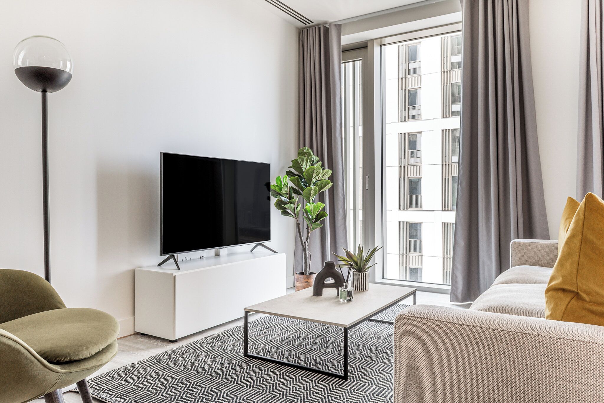 A modern living room with a flat-screen TV on a white cabinet, a coffee table, a sofa, a floor lamp, and a large window with curtains.