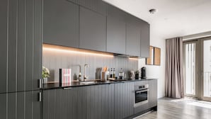 A modern kitchen with dark cabinetry, a built-in oven, and a countertop with various appliances and decor items. - Urban Rest Stratford (London)