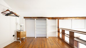 Una habitación con suelo de madera, un armario blanco con estantes abiertos, un banco de madera y una percha con ropa.