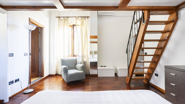 Una habitación con suelo de madera, un sillón gris, una cama blanca, una escalera de madera y una pequeña mesa blanca.