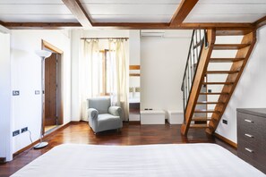 Una habitación con suelo de madera, un sillón gris, una cama blanca, una escalera de madera y una pequeña mesa blanca.