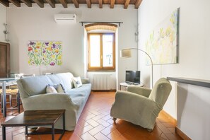 Una sala de estar con sofá, sillón y una mesita. Se ve una ventana con vistas, un radiador y un televisor sobre un mueble.