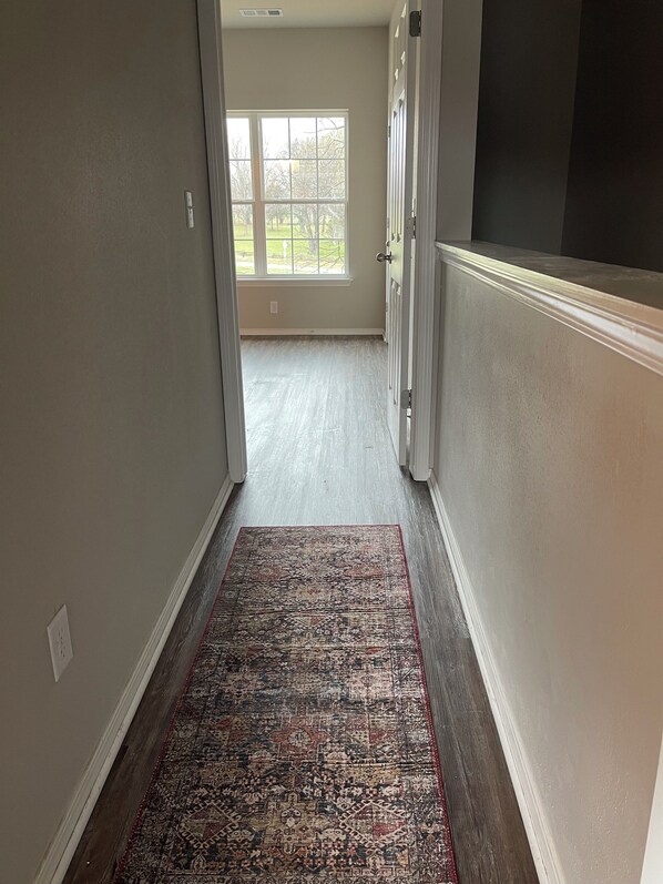 A hallway with a patterned rug, a window with a view of trees, and a door with a light switch on the wall.