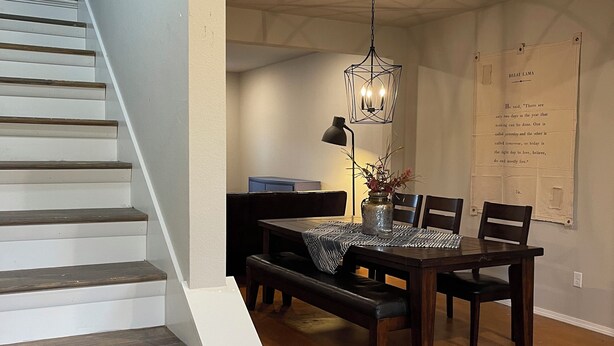 Un escalier menant à une salle à manger où une table est dressée pour quatre personnes, avec un luminaire suspendu et un mur orné d’une citation encadrée.