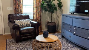 Un salon avec un canapé en cuir marron, un tapis à motifs, une table basse ronde en bois, une commode bleue et un téléviseur fixé au mur.