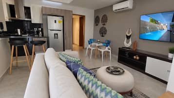 A modern living room with a sofa, a dining area with chairs, a kitchen with a refrigerator, and a TV mounted on the wall.