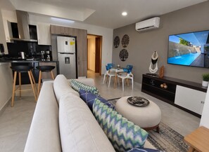 A modern living room with a sofa, a dining area with chairs, a kitchen with a refrigerator, and a TV mounted on the wall.