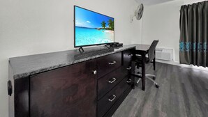 Una habitación con televisión mostrando una escena de playa, un escritorio con una silla, un aparador de madera oscura y un ventilador de techo.