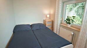 A small, single-bed room with a window, a bedside table with a lamp, and a potted plant.