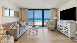 A living room with a grey sofa, a round coffee table, a patterned armchair, a TV on a white cabinet, and a balcony with a view of the beach. - Hart of the Sea: 3rd floor, remodeled, beachfront, stylish, incredible amenities (Panama City Beach)
