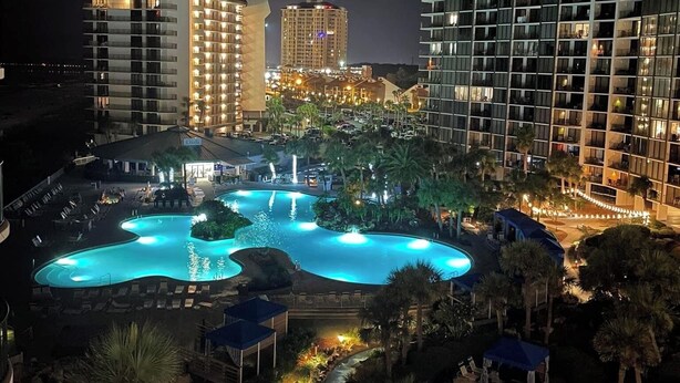 A nighttime view of a hotel with a large, illuminated swimming pool surrounded by palm trees and lounge chairs. - Hart of the Sea: 3rd floor, remodeled, beachfront, stylish, incredible amenities (Panama City Beach)